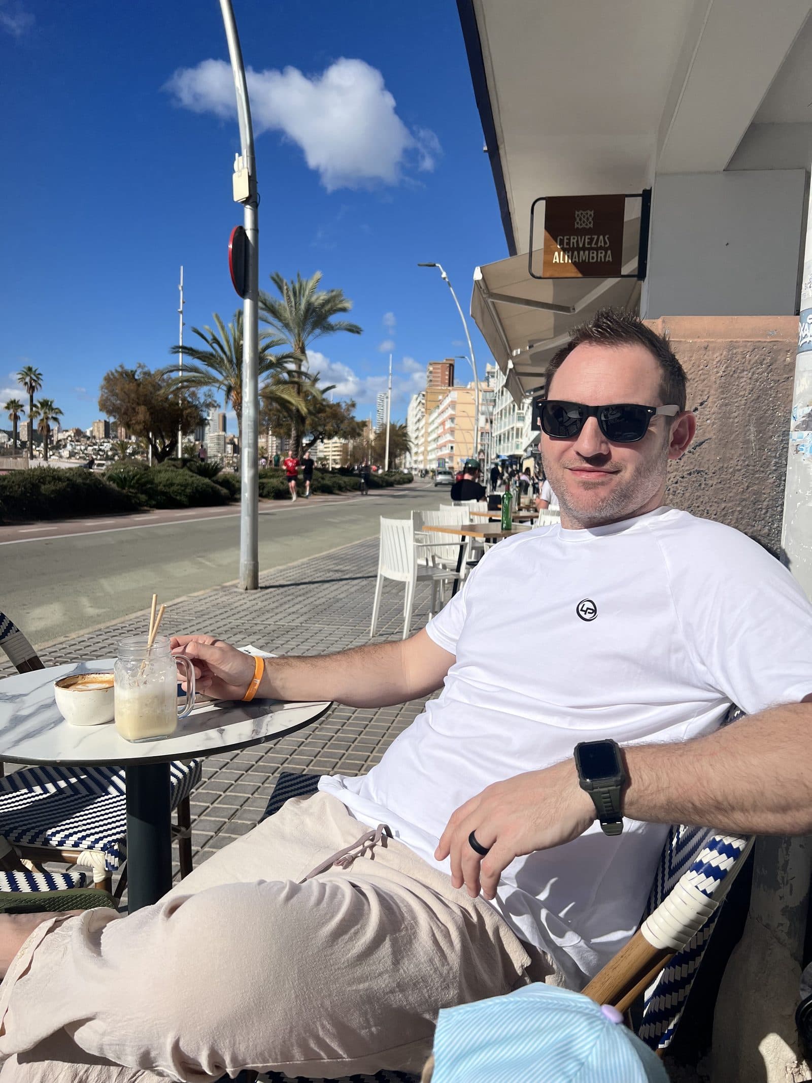 Liam relaxing at a beachfront cafe on Benidorm promenade with a smoothie, palm trees and the Poniente skyline behind