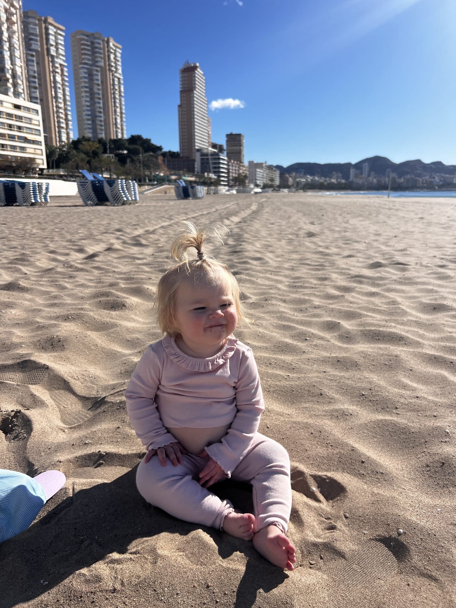 Tallulah sitting on Poniente Beach in Benidorm, smiling at the camera with high-rise buildings behind her