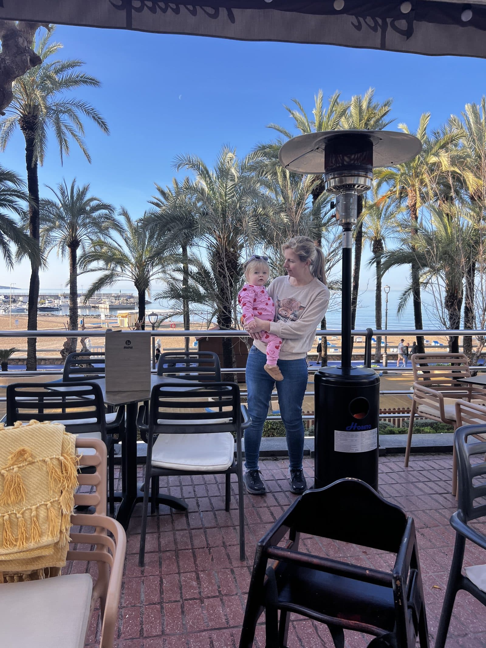 Leila holding Tallulah at a sunny outdoor cafe terrace in Benidorm with palm trees