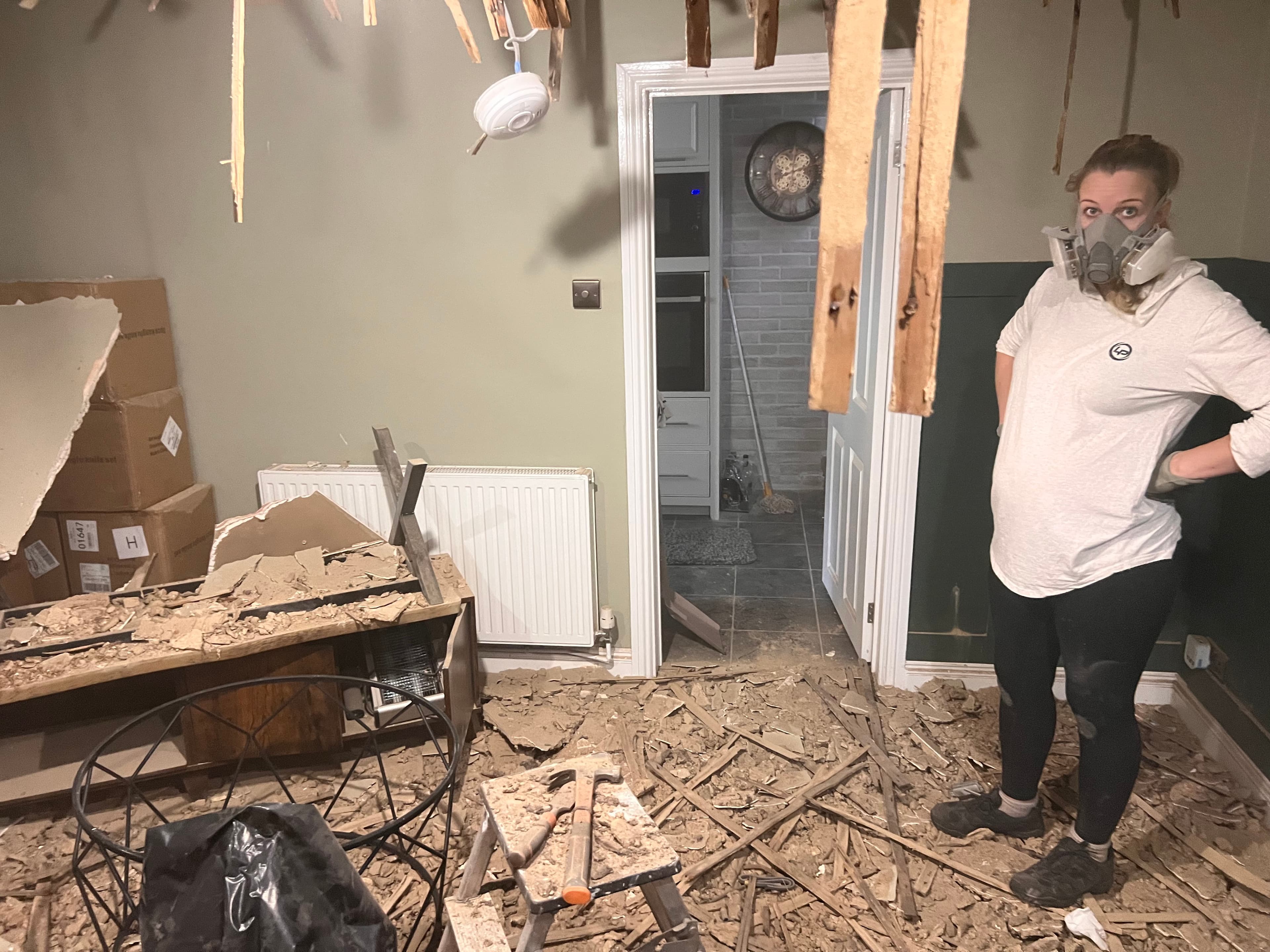 Leila standing in the living room with the ceiling collapsed around her, pregnant, wearing a gas mask during renovations