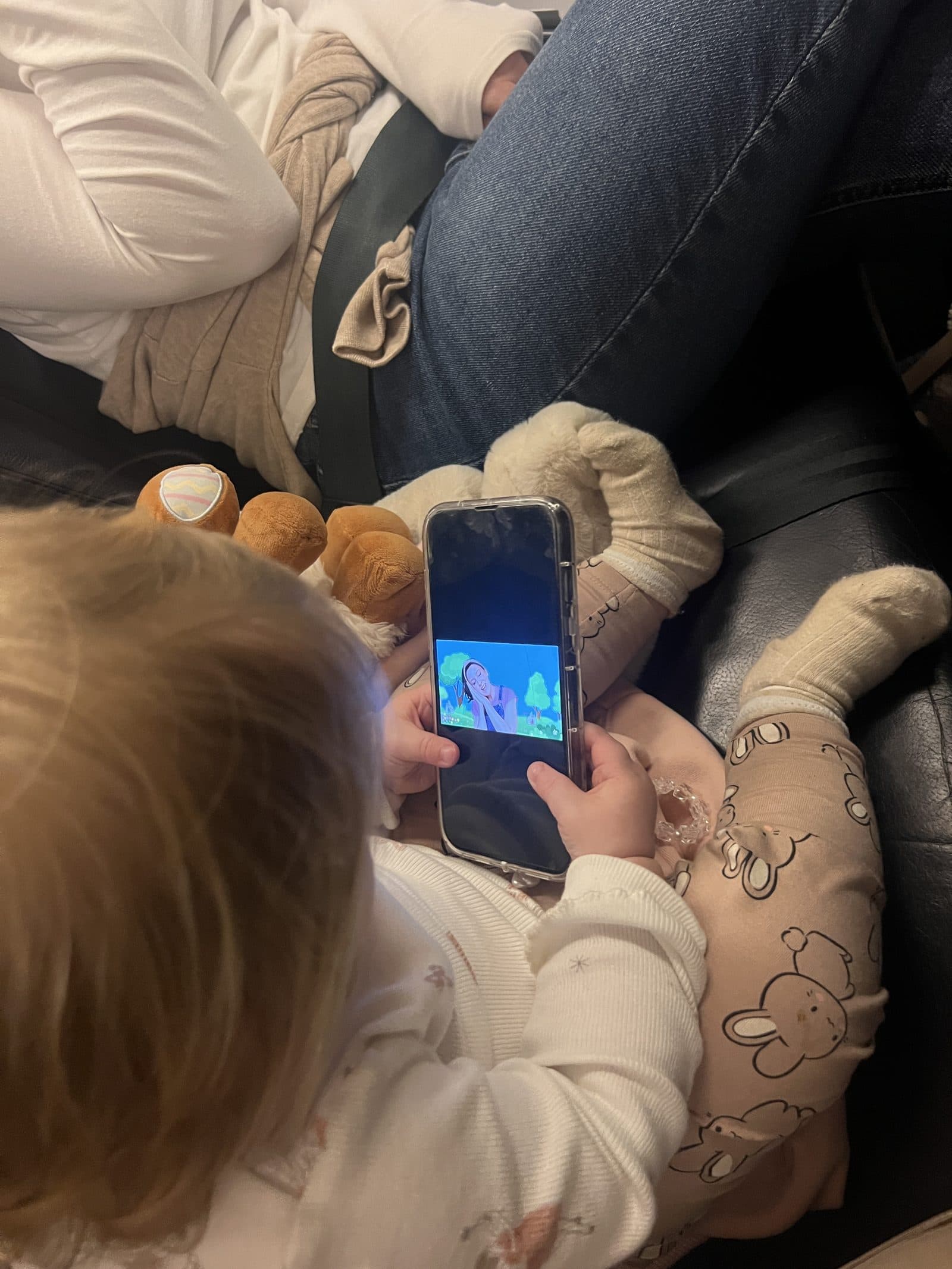 Baby watching cartoons on a phone during the Ryanair flight, lying on mum's lap with a dummy and cuddly toy