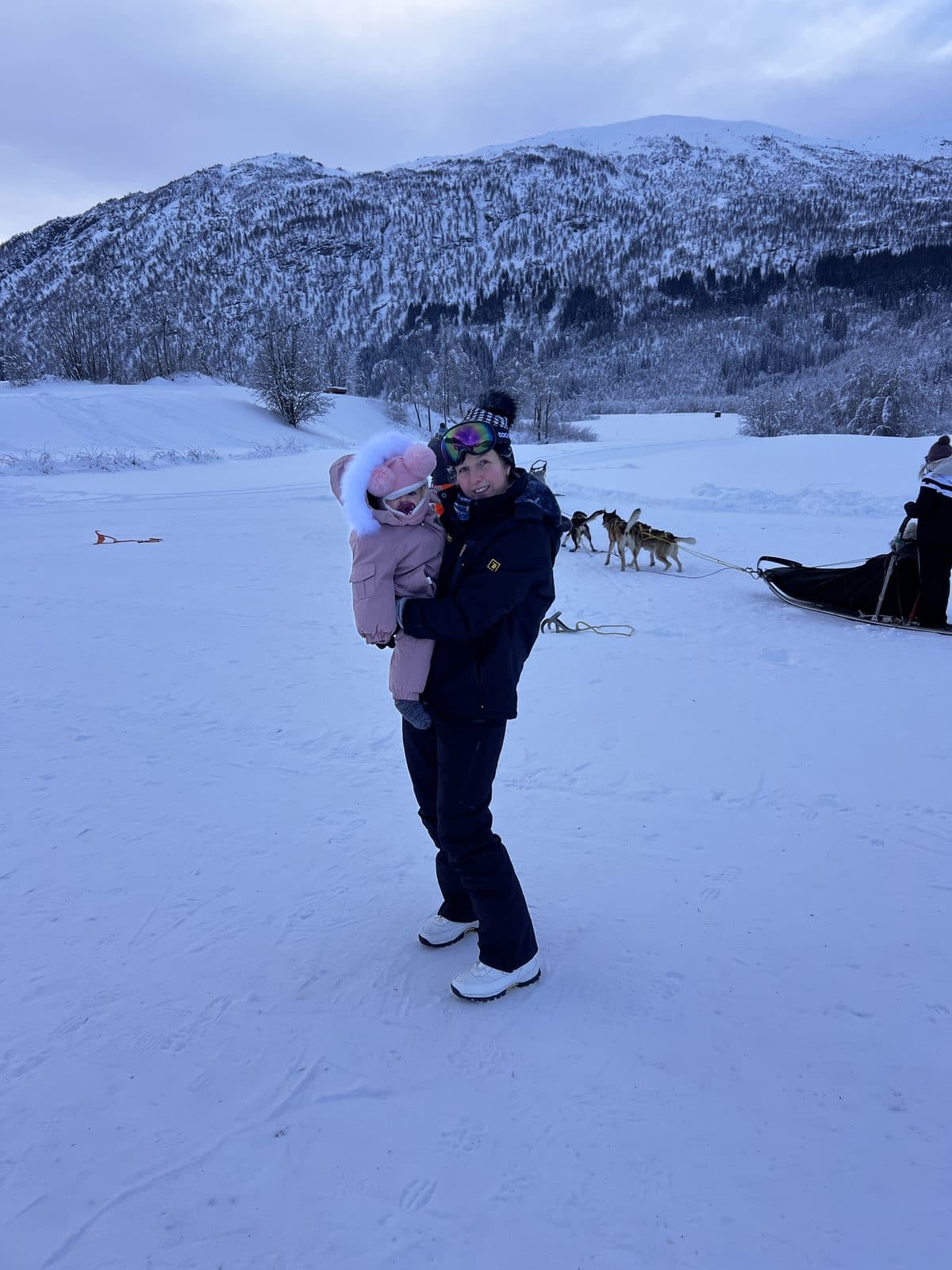 Mum holding baby in the snow at Myrkdalen with husky sled dogs and Norwegian mountains behind