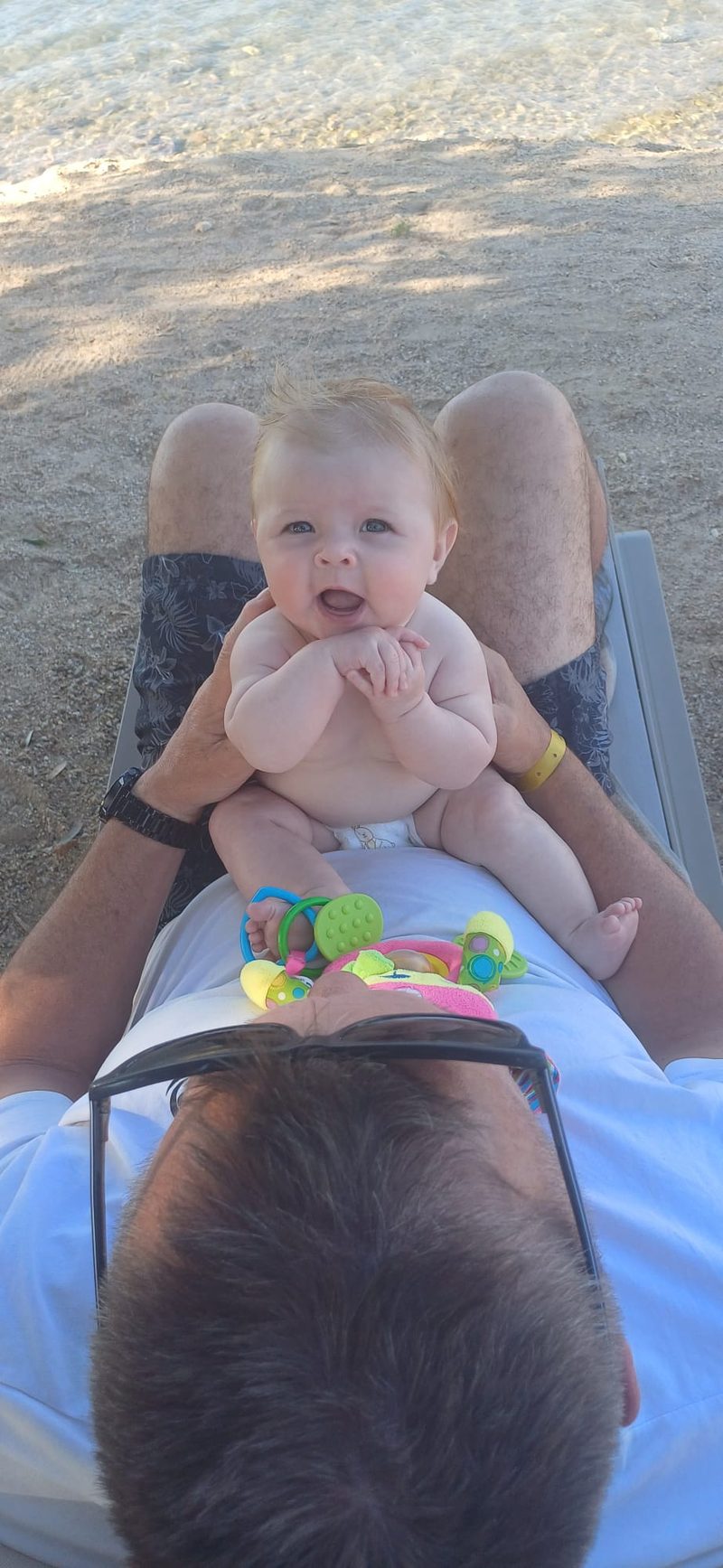 Happy 4-month-old baby sitting on dad's legs at the beach in Akbuk Turkey looking at camera